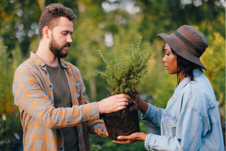Two people outdoors exchanging a potted plant, holding the root ball together in a garden setting surrounded by greenery.